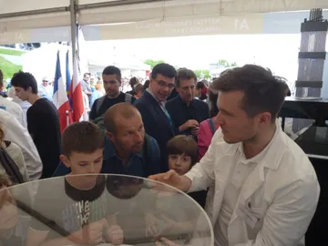 Osoba w białym fartuchu objaśnia dzieciom makietę reaktora; w tle francuskie flagi