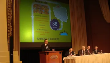 Prelegent przy mównicy przedstawia slajd o grupach docelowych Atomowego Autobusu; obok panel przy stole