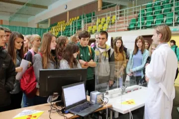 Osoba w białym fartuchu pokazuje uczniom doświadczenie przy laptopie