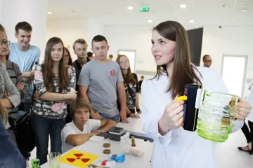 Osoba w białym fartuchu pokazuje uczniom miernik przy stole z eksponatami