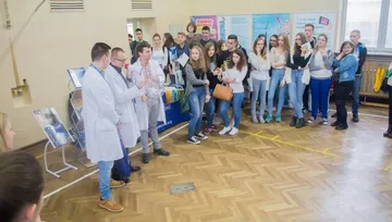 Osoby w białych fartuchach prowadzą pokaz dla uczniów w sali gimnastycznej