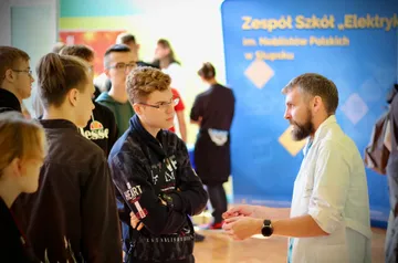 Osoba w białym fartuchu rozmawia z uczniami przy roll-upie Zespołu Szkół „Elektryk”