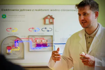 Osoba w białym fartuchu objaśnia planszę o elektrowni jądrowej z reaktorem wodnym ciśnieniowym