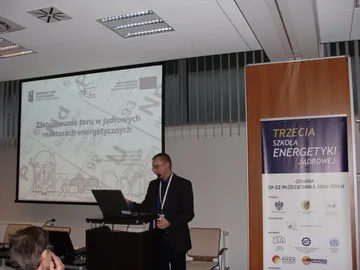 Osoba przy mównicy; na ekranie slajd „Zastosowanie toru w jądrowych reaktorach energetycznych”, obok roll-up Trzeciej Szkoły Energetyki Jądrowej