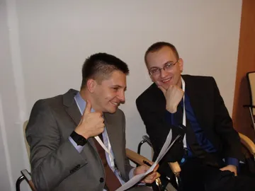 Dwie osoby z identyfikatorami siedzą na sali; jedna pokazuje uniesiony kciuk