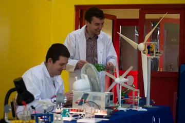 Osoba w białym fartuchu pokazuje uczniom model turbiny wiatrowej i doświadczenia