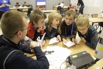 Dzieci pochylają się nad stołem z kartkami i miernikiem; obok osoba prowadząca zajęcia