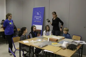 Dzieci przy stole z eksponatami; obok roll-up Uniwersytetu Dzieci i dwie osoby prowadzące zajęcia