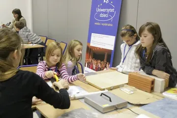 Dzieci wykonują pomiary przy stole z próbkami; w tle roll-up Uniwersytetu Dzieci