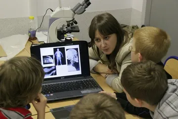 Dzieci oglądają na laptopie zdjęcia rentgenowskie; obok mikroskop i osoba prowadząca zajęcia