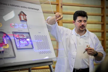 Osoba w białym fartuchu objaśnia planszę z modelem elektrowni