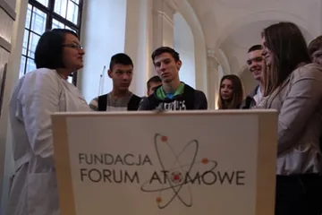 Osoba w białym fartuchu rozmawia z uczniami przy mównicy z logo Fundacji
