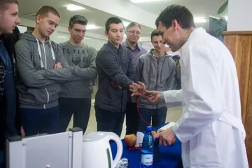 Osoba w białym fartuchu pokazuje uczniom doświadczenie przy stole