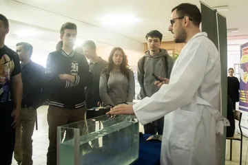 Osoba w białym fartuchu prowadzi doświadczenie z akwarium przed uczniami