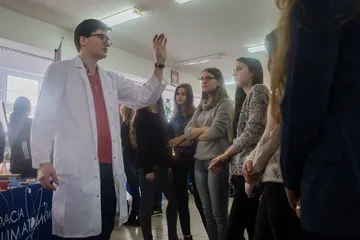 Osoba w białym fartuchu z uniesioną ręką opowiada uczniom