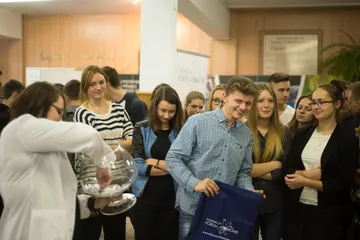 Osoba w białym fartuchu z kulą wypełnioną kolorowymi kulkami przed uczniami